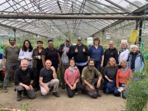 Sixteen men and women smiling to camera in a group photo taken in the glasshouse at Hawkwood Plant Nursery.