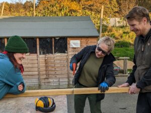 Two people stand on either side a length of timber to steady it, while a third person saws through it.