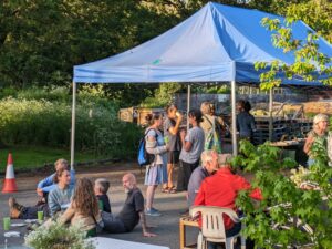 Groups of people standing and sitting chatting to each other close to a small marquee and surrounded by trees in summer