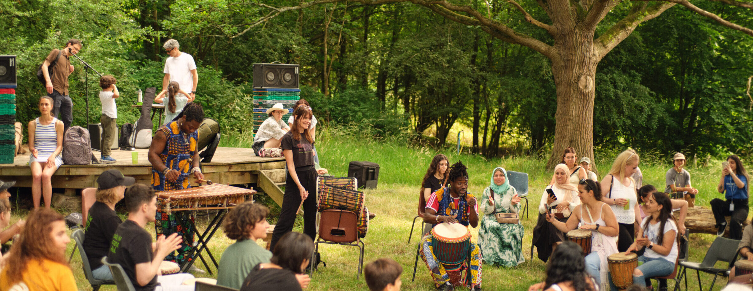 Celebratory gathering of people, some playing the drums, in a woodland clearing at Hawkwood Plant Nursery