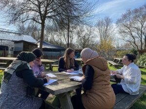 Five women sit studying at a wooden picnic table surrounded by greenery and trees.